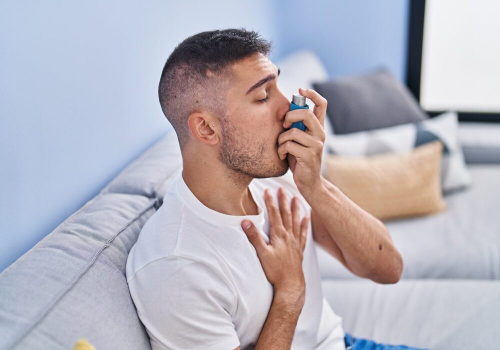 Young hispanic man using inhaler sitting on sofa at home